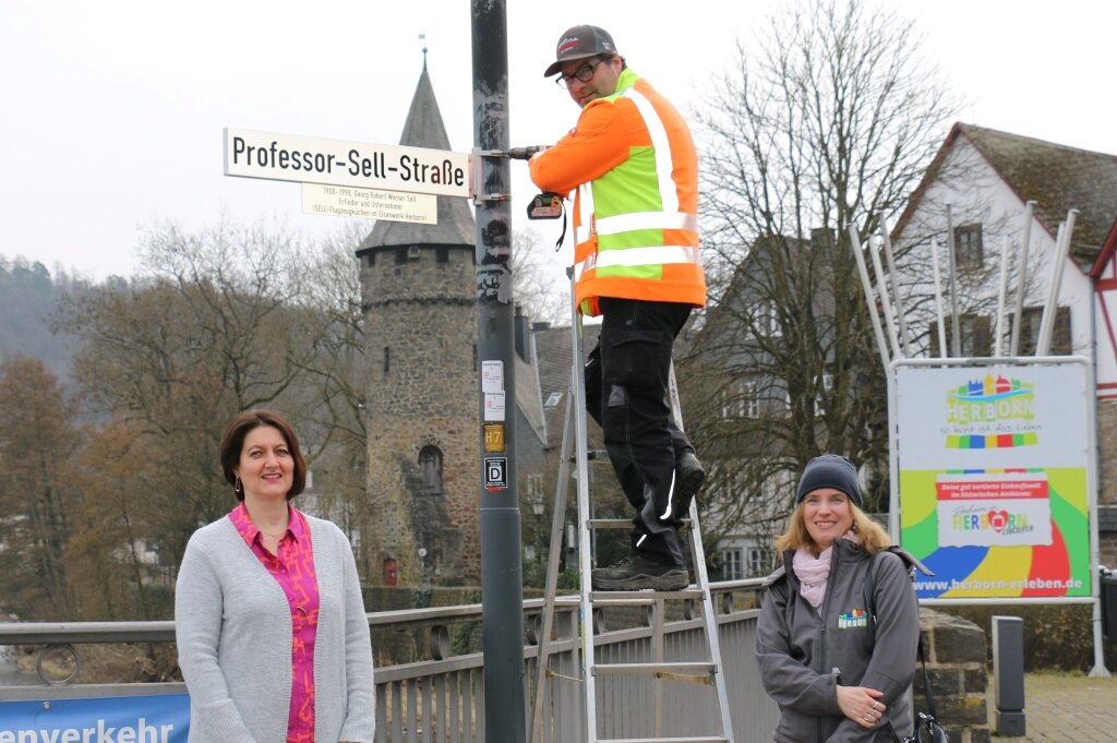 Zwei Frauen und ein Mann auf einer Leiter. Neue Zusatzschilder erklären die Straßennamen.