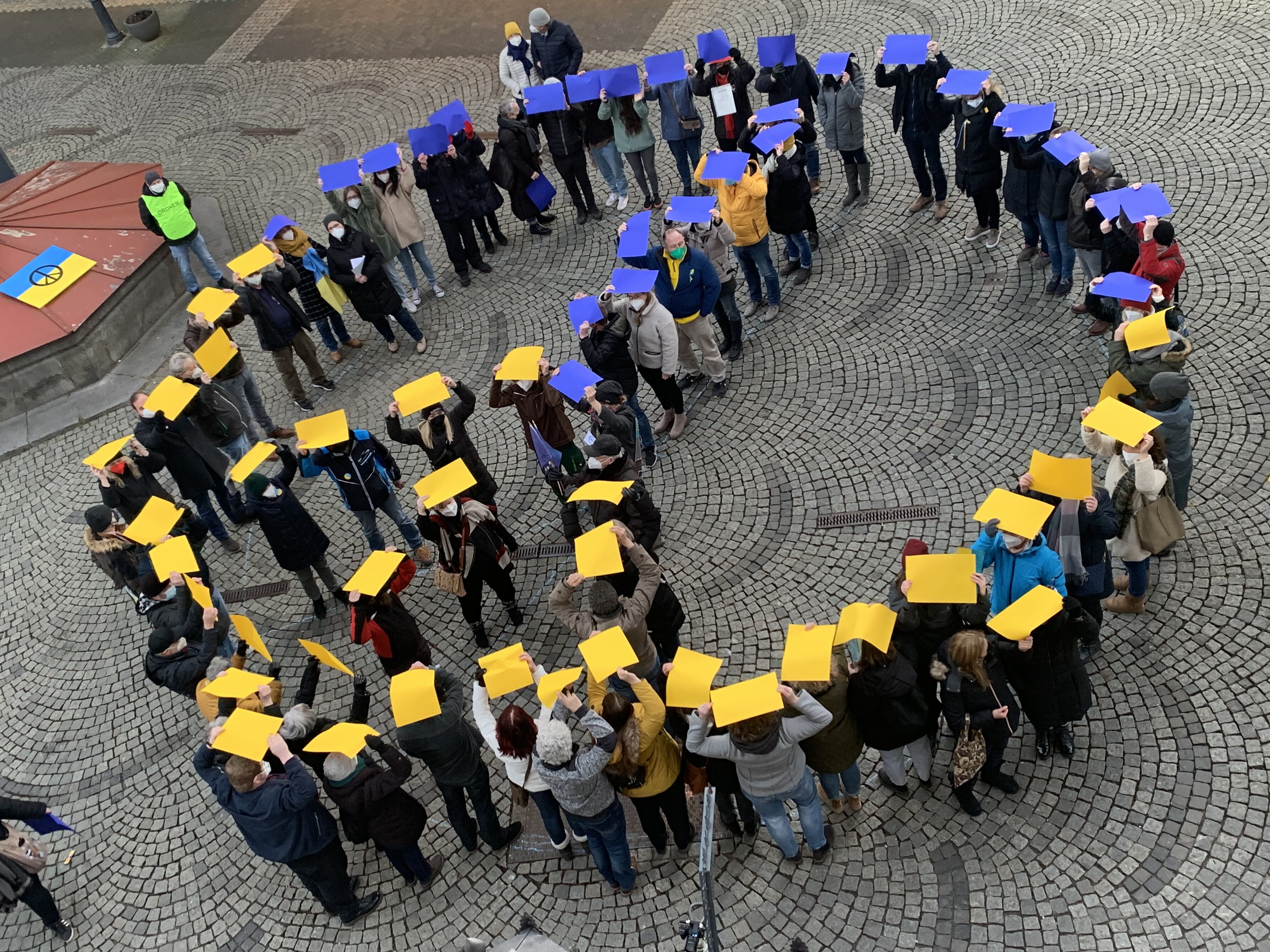 Peace-Zeichen mit Menschen, Menschen halten gelb-blaue Zettel als Zeichen der Solidarität mit der Ukraine empor.