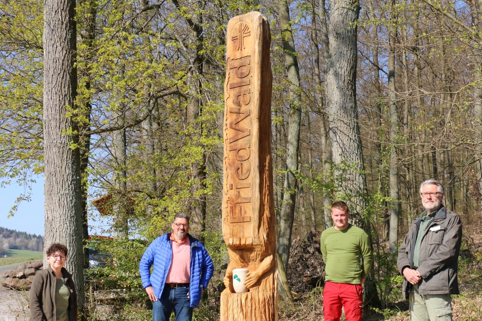 Eine Frau und drei Männer stehen an einer Urnenstele im Wald.